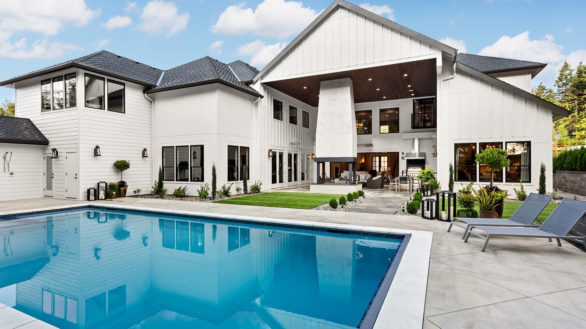 A modern white house features a large swimming pool, lush lawn, and outdoor seating area under a covered patio with fireplace.