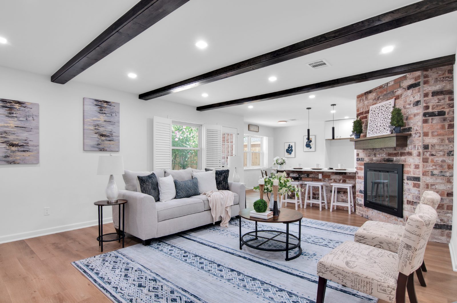 Open living room with dark ceiling beams, a brick fireplace and a kitchen bar