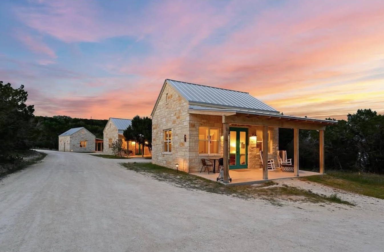 Warm lights shine from limestone casitas along a quiet Hill Country drive beneath a pink and gold sunset.