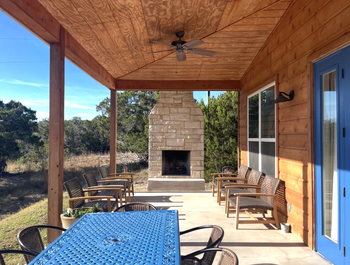 A sunny covered porch at Hill Country Retreats with chairs gathered around a limestone fireplace and a blue table.