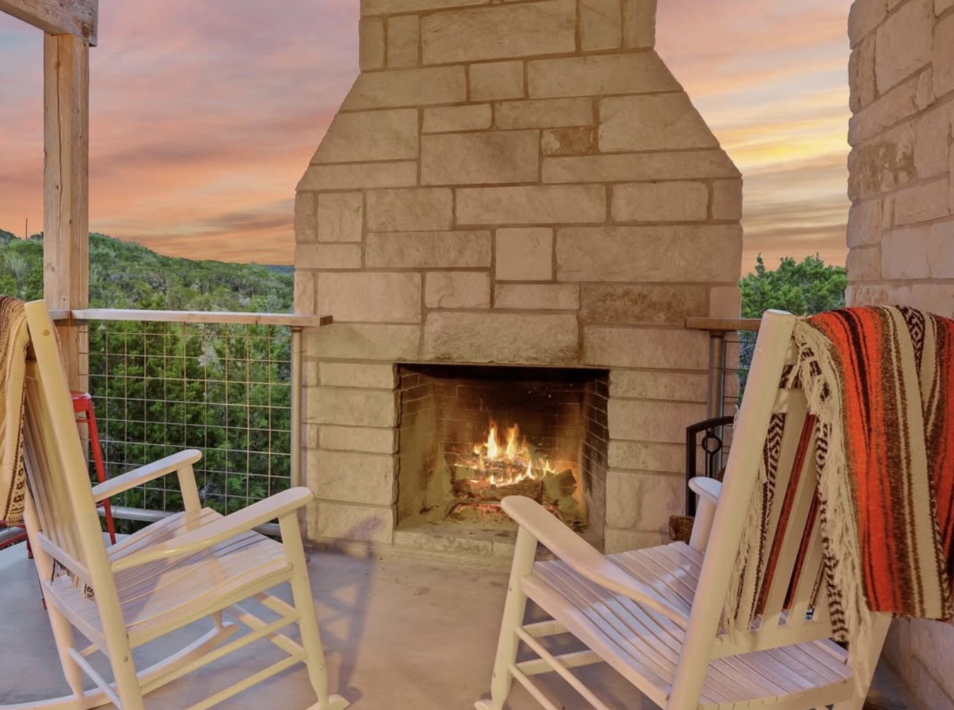 Two rocking chairs with blankets beside a glowing limestone fireplace, overlooking the Hill Country at sunset.