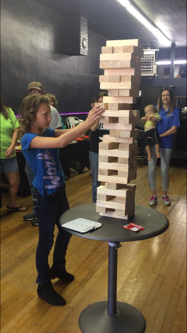 Guest removes a wooden block from a giant stacking game while others watch the challenge.