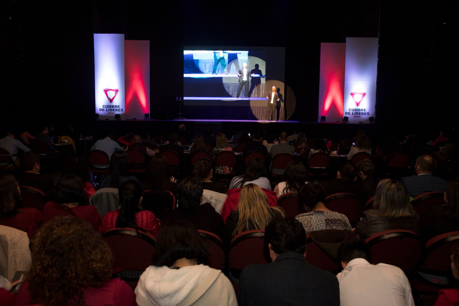 Sala completa de la Cumbre vista desde atrás, con el escenario y las pantallas al fondo.