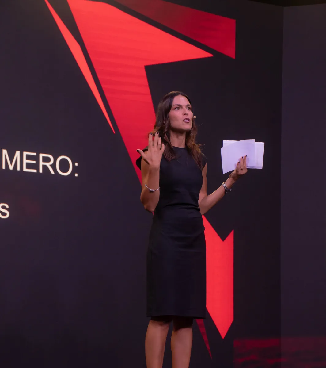 Ponente de vestido negro hablando de pie en el escenario, con notas en la mano.