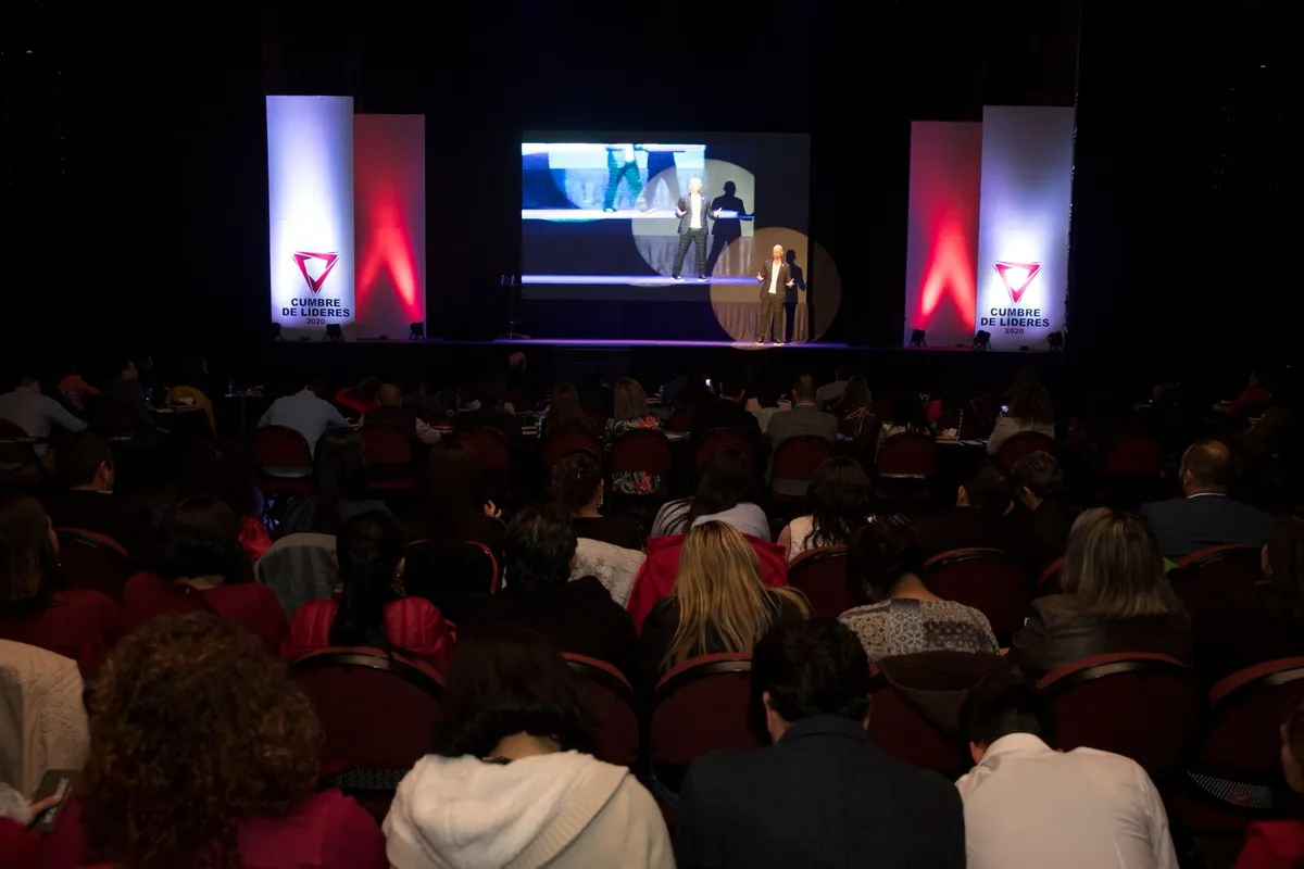 Sala completa de la Cumbre vista desde atrás, con el escenario y las pantallas al fondo.