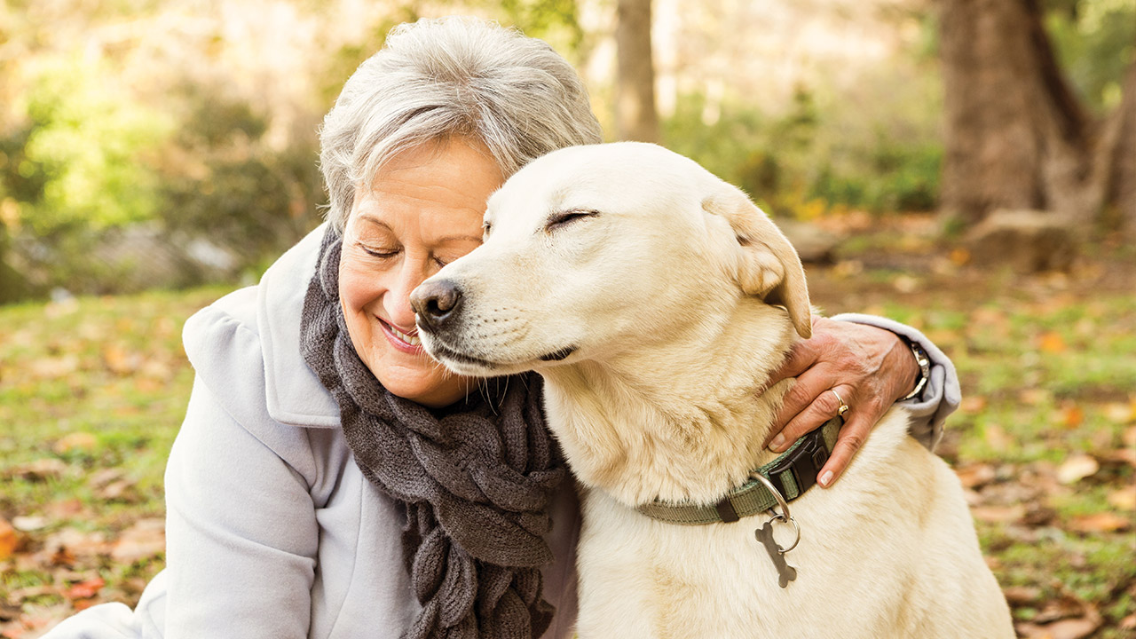 A woman outdoors in autumn with her arm around a yellow Labrador retriever.