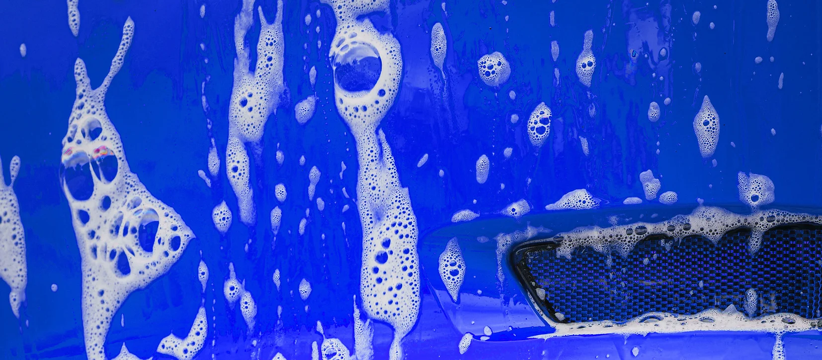 Close-up of thick soap suds on blue vehicle surface during hand wash detailing.