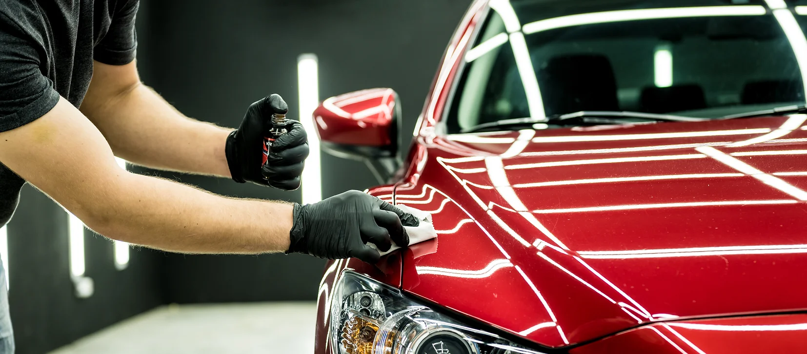 Detailer applying ceramic coating to red car with an applicator pad and protective gloves under detailing lights.