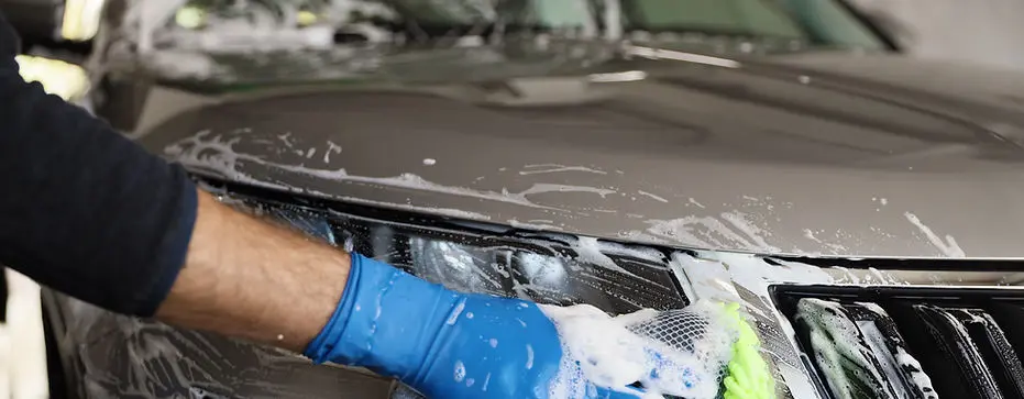 Auto detailer using a microfiber mitt and blue glove to hand wash a vehicle’s front grille and hood.