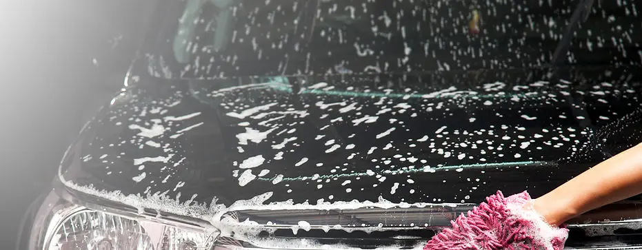 Close-up of a black car hood being hand-washed with pink microfiber mitt and thick foam suds.