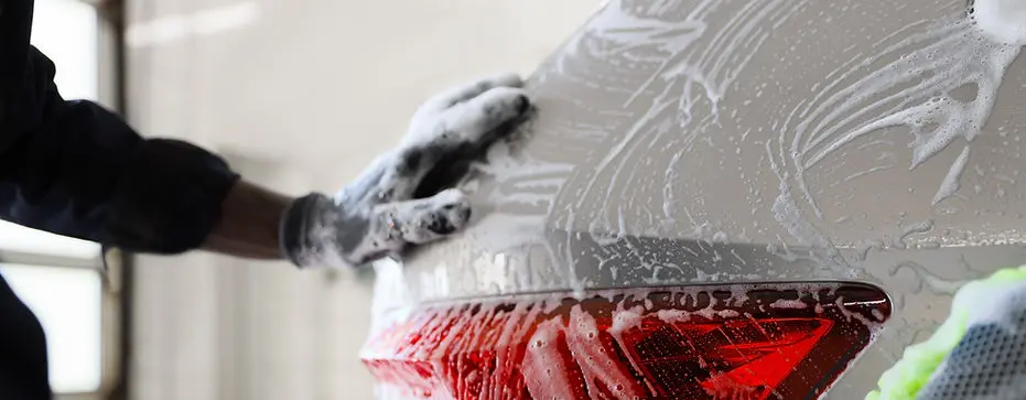 Detailer hand-washing a car’s rear panel with soap and a microfiber mitt.