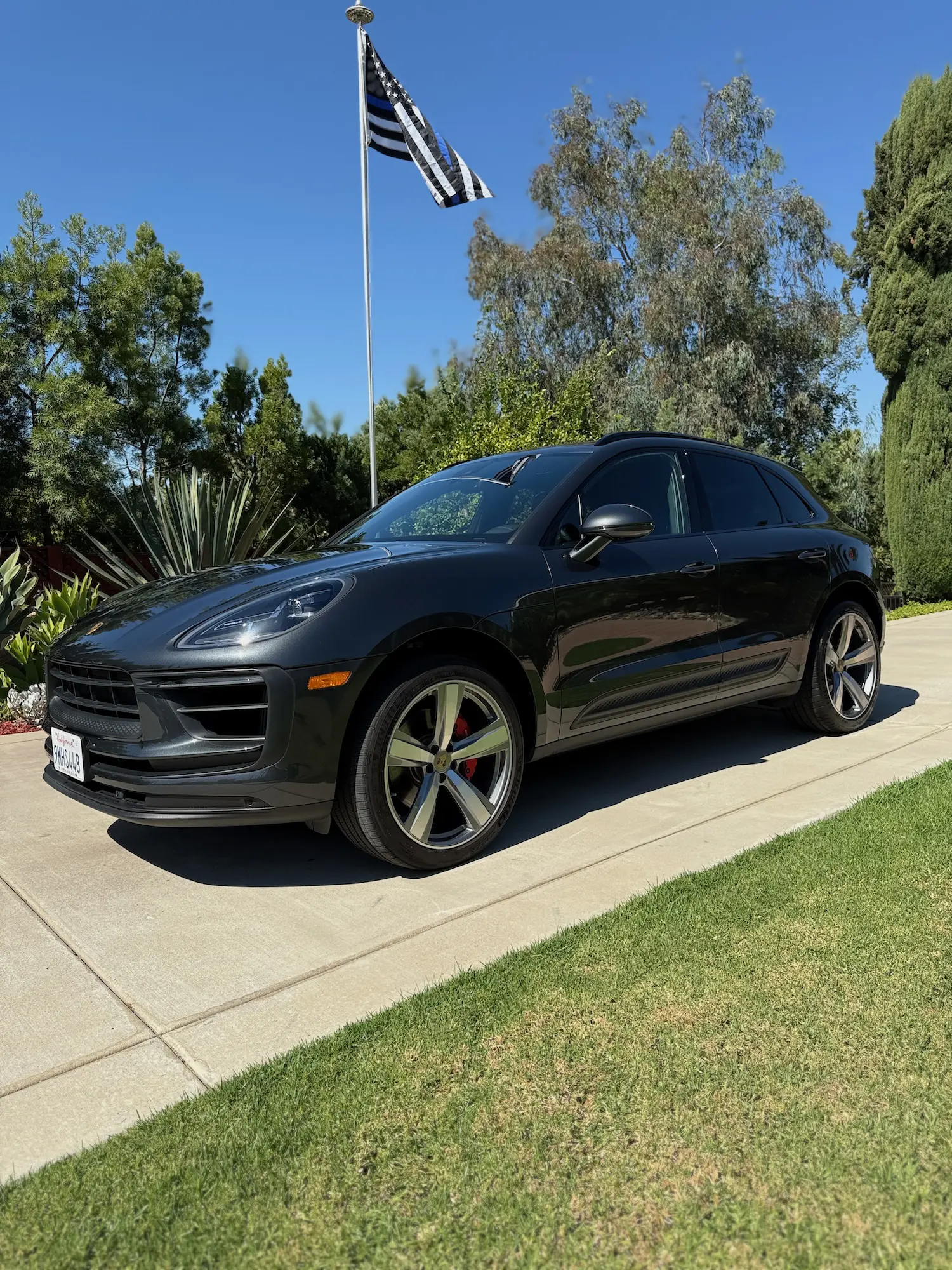 Black Porsche Macan parked on a sunny driveway with lush greenery and a flag in the background