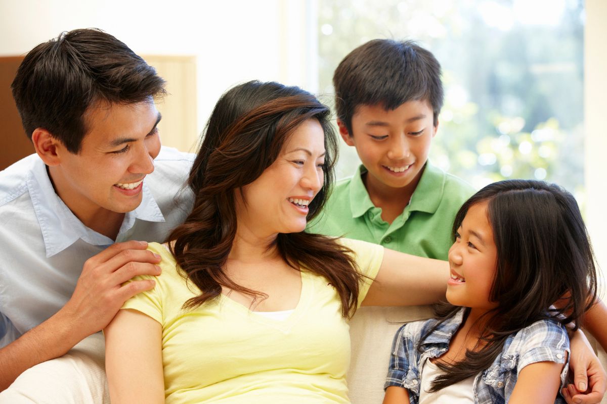 Parents, with their young son and daughter, sit on a couch, smiling and chatting while they teach their kids Chinese at home.