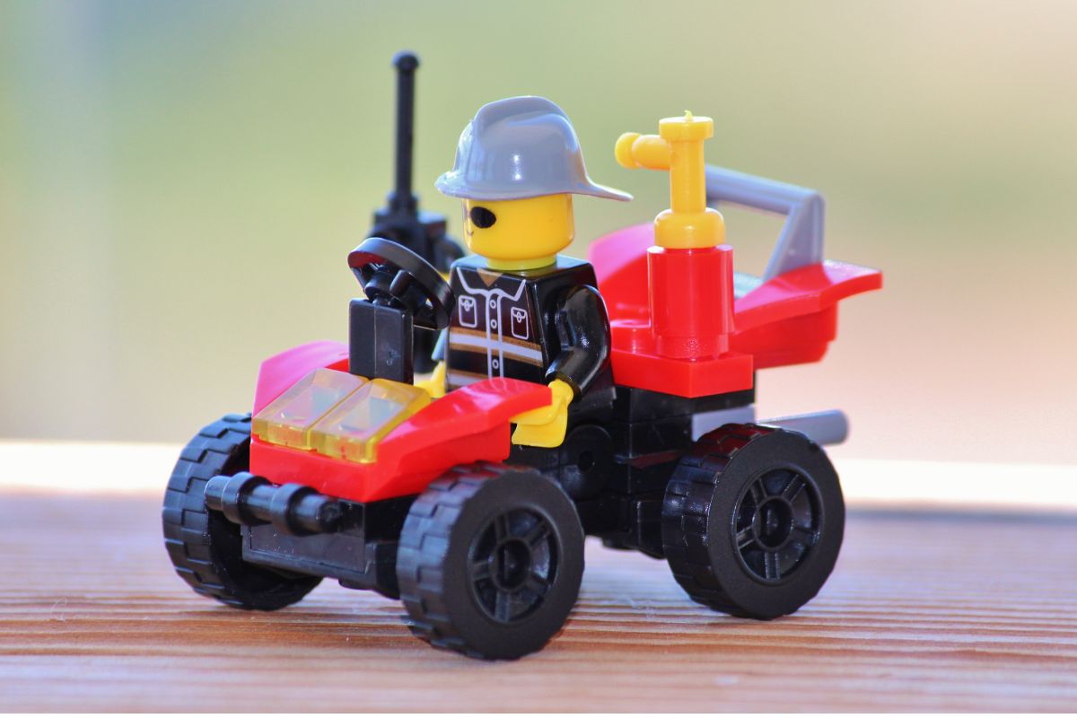 Lego firefighter figure in a grey hat drives a small red fire response vehicle with black wheels and a rear mounted water pump on a wooden surface.
