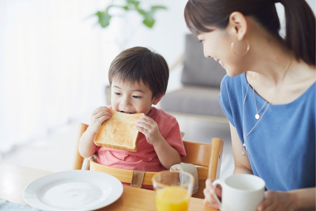 Mum smiles at toddler in a high chair eating toast at the breakfast table with a mug and glass of orange juice.