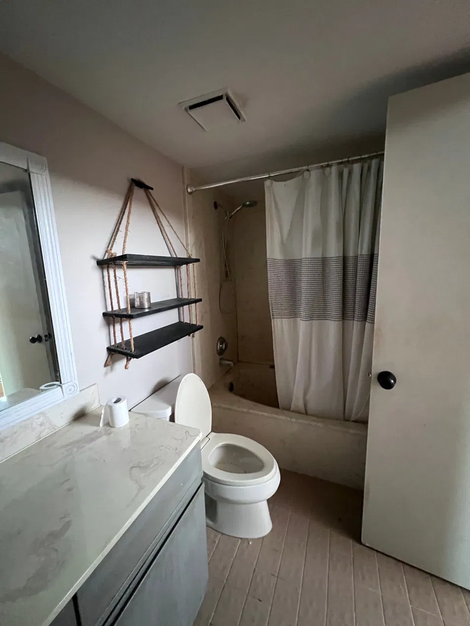 Before: dated master bathroom with a shower curtain over the tub, worn vanity top and dated flooring