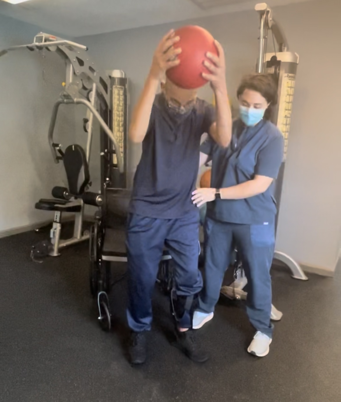 Patient holding a red medicine ball overhead with therapist spotting in a private fitness session.