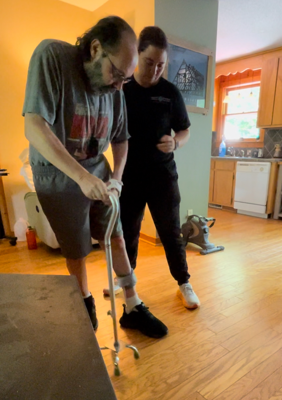 A Neuro Nation therapist supporting a patient practicing standing with a walker in his living room.