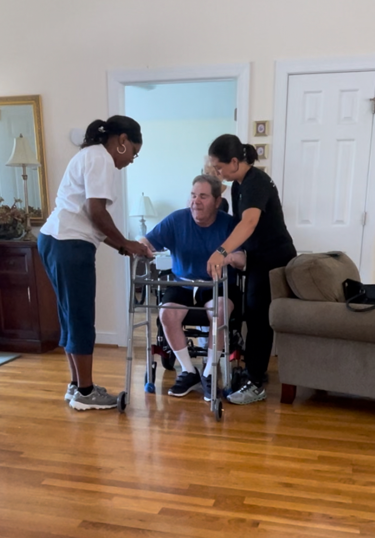 Two therapists supporting a seated patient as he stands with a walker during an in-home private consultation.
