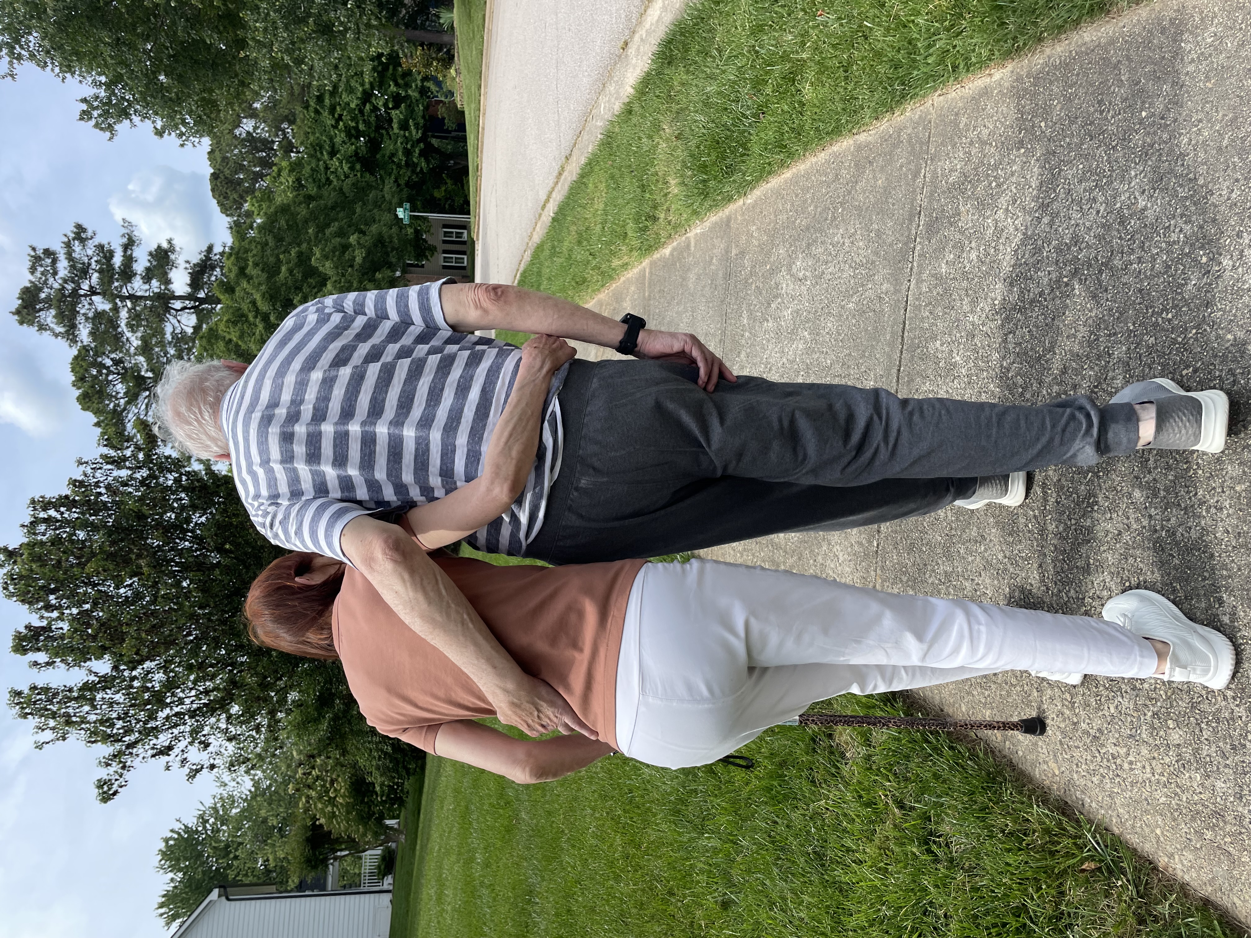 Therapist and patient walking together outdoors on a residential sidewalk, arms supporting one another.