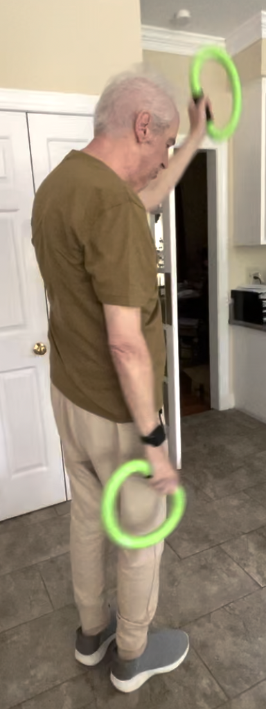 Older man performing a PWR!Moves standing exercise with resistance rings during in-home Parkinson's therapy.