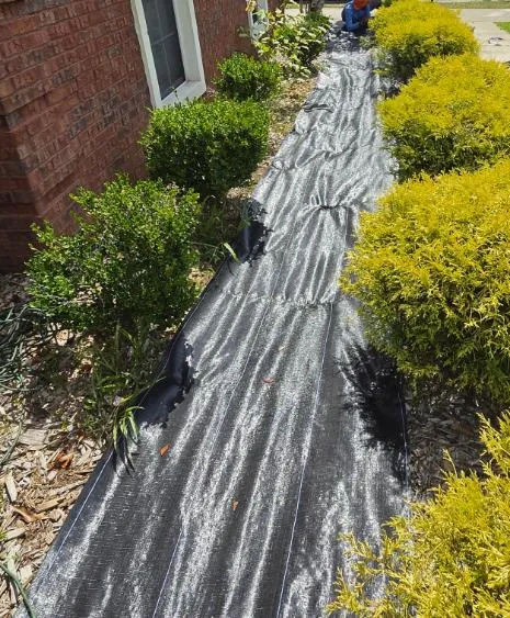 Black landscape fabric laid along a narrow bed beside a brick wall with a crew member working at the far end