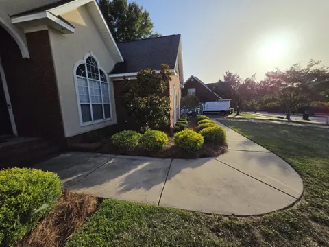 Curving concrete walkway beside a pine straw mulch bed with small trimmed shrubs and a brick and stucco house