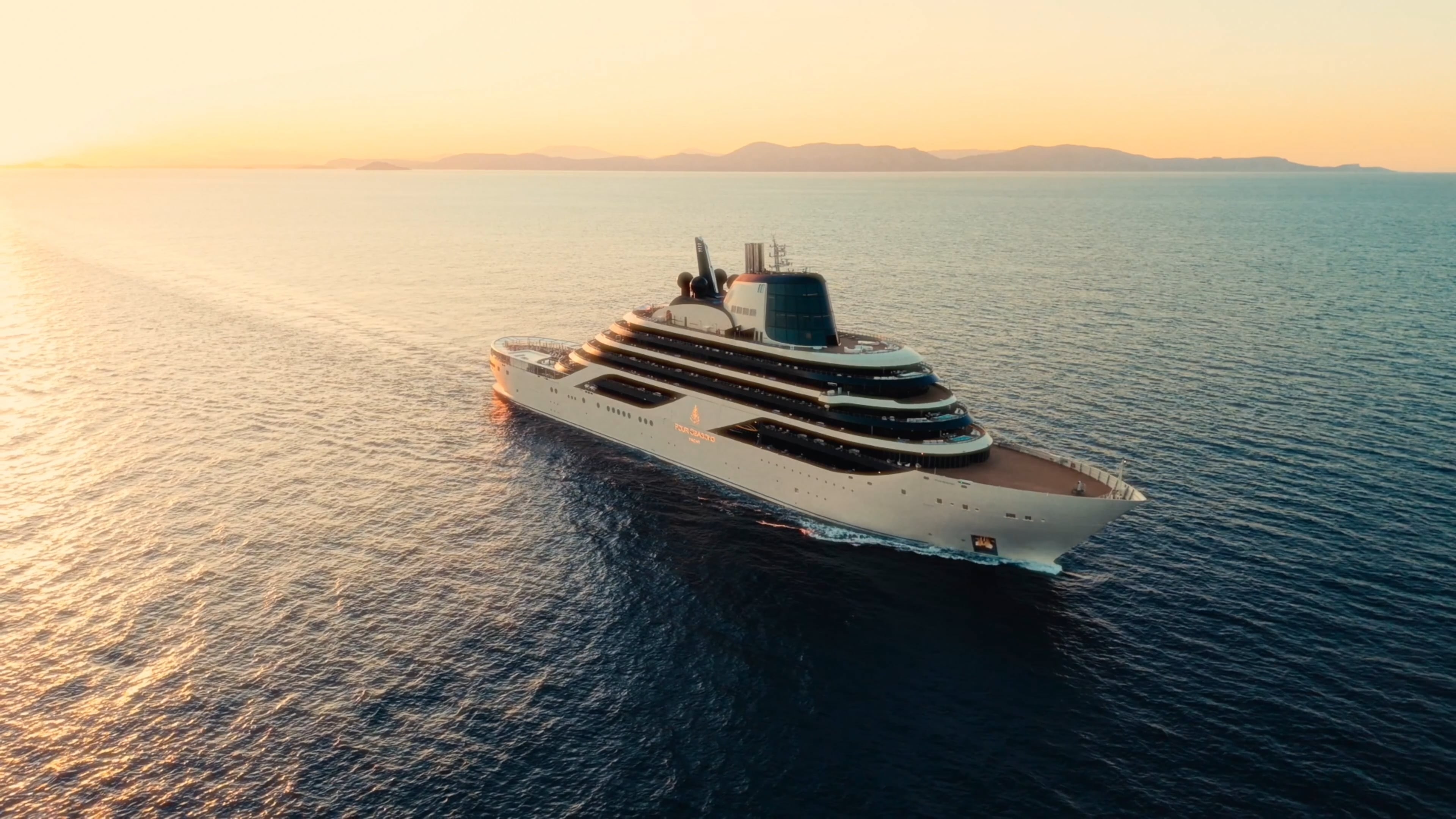 Four Seasons I under way at sunset on open water, her white hull and tiered aft decks catching low golden light with a distant mountainous coastline on the horizon