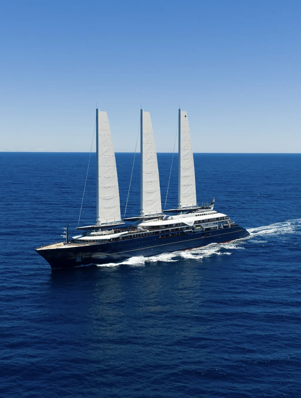 The Orient Express Corinthian under way on deep blue open sea, carrying three tall white rigid solid sails above a dark navy hull