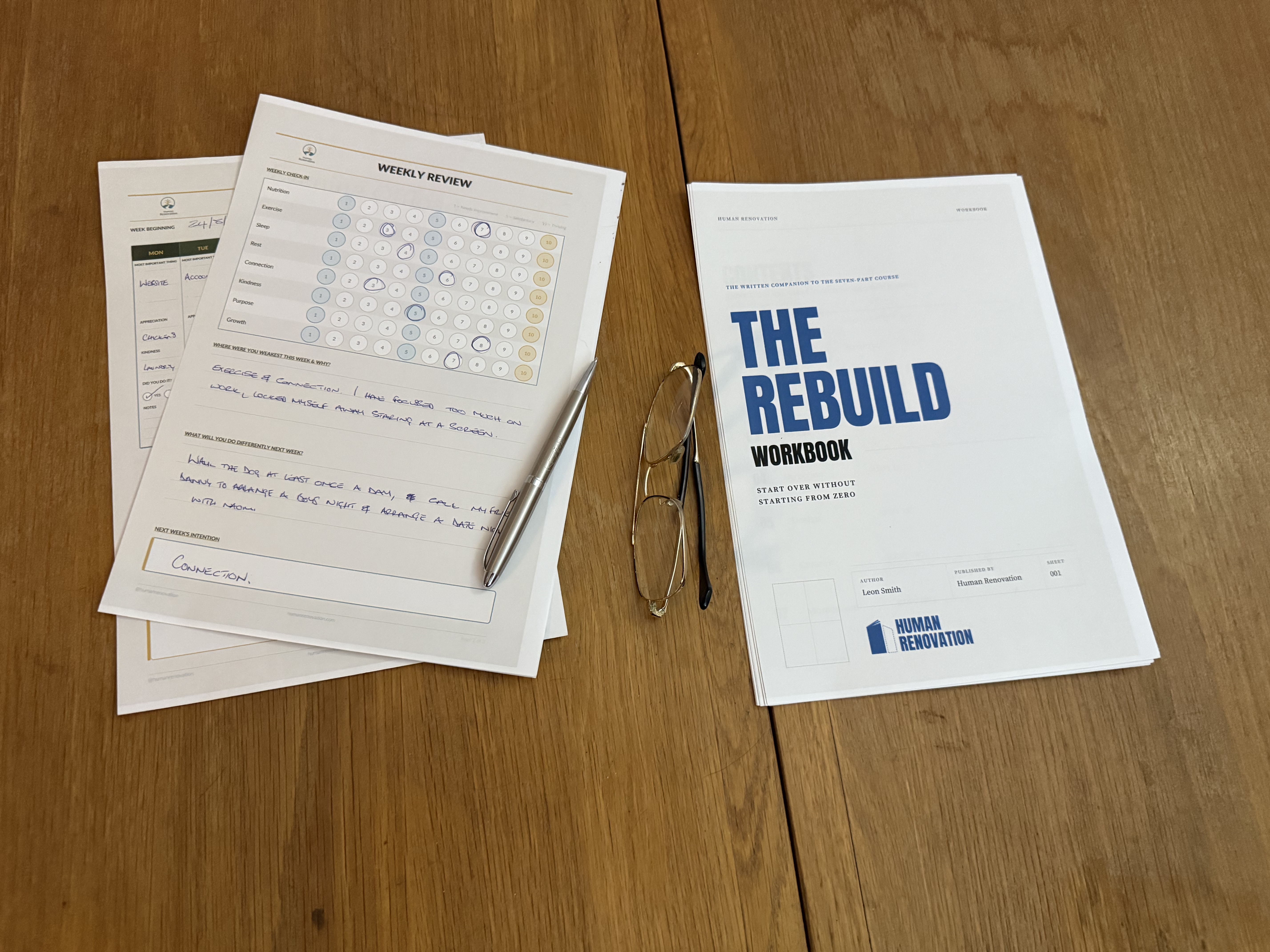A Weekly Review sheet filled in by hand beside the workbook, pen and reading glasses on the table