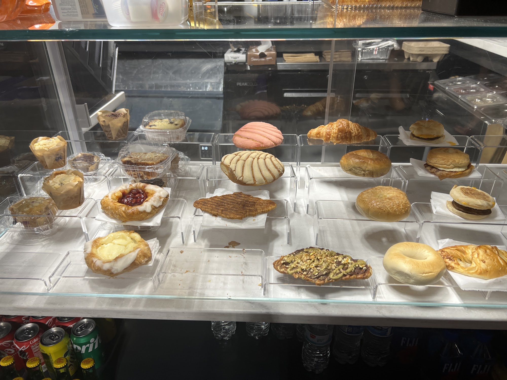 Pastry case close-up — conchas, danishes, flat croissant with pistachio, bagels, English muffins