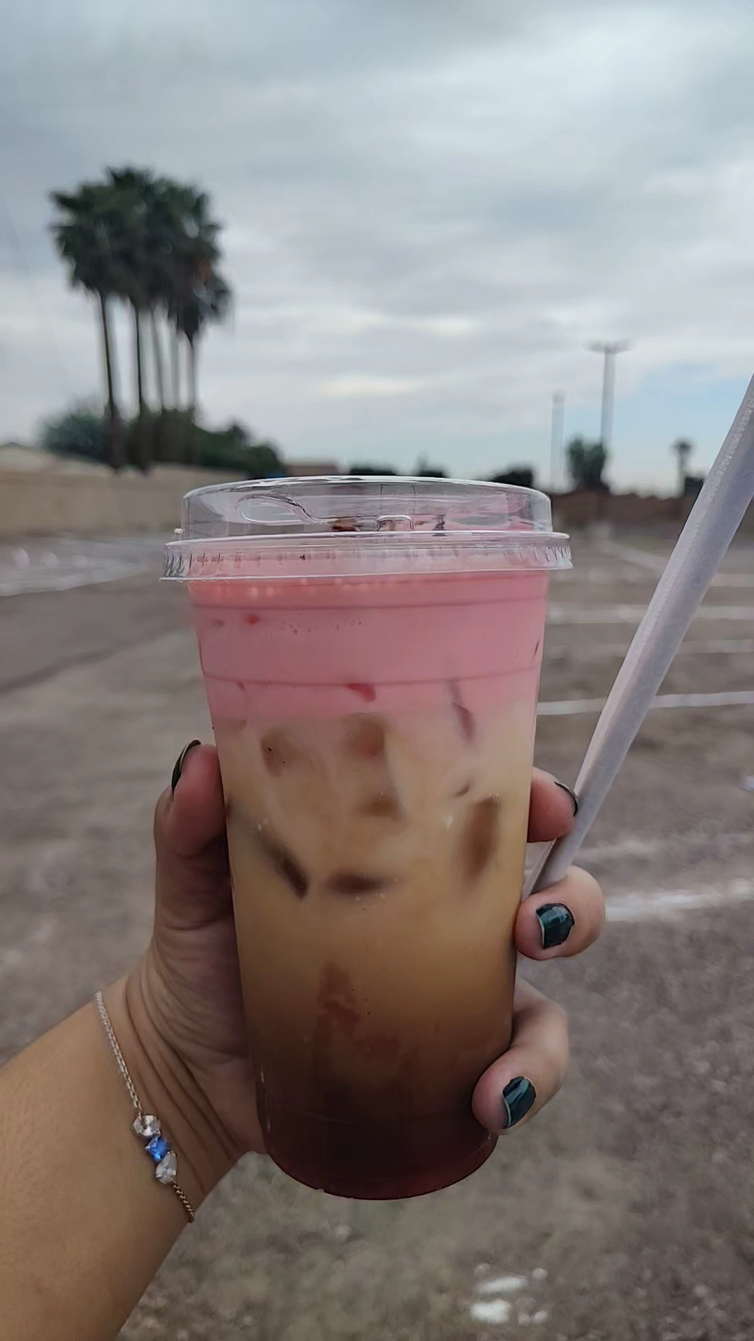 Pink strawberry cold foam latte held outdoors with palm trees and cloudy sky behind