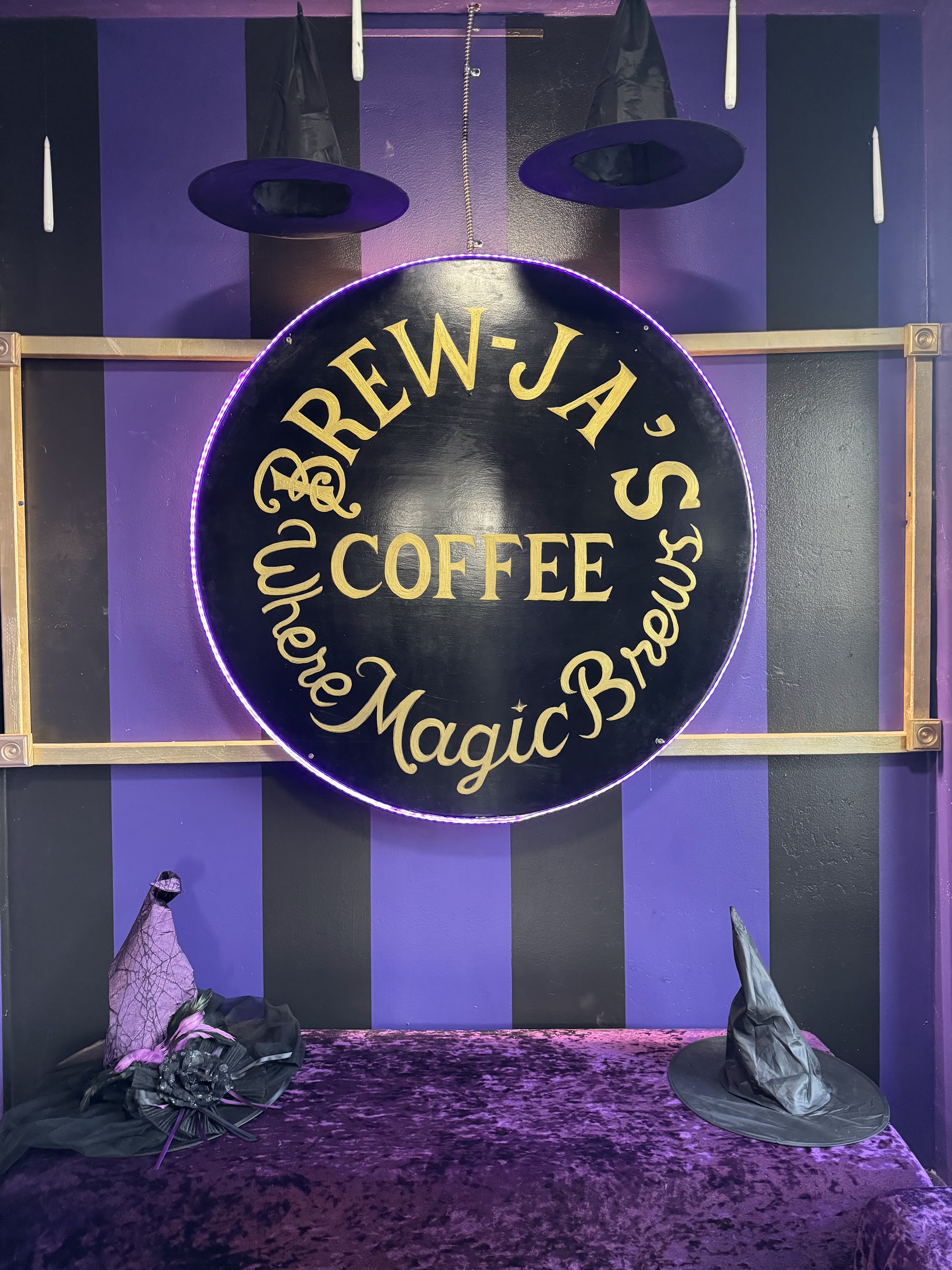 Round Brew-Ja's Coffee neon sign — gold on black, purple and black striped wall, witch hats hanging