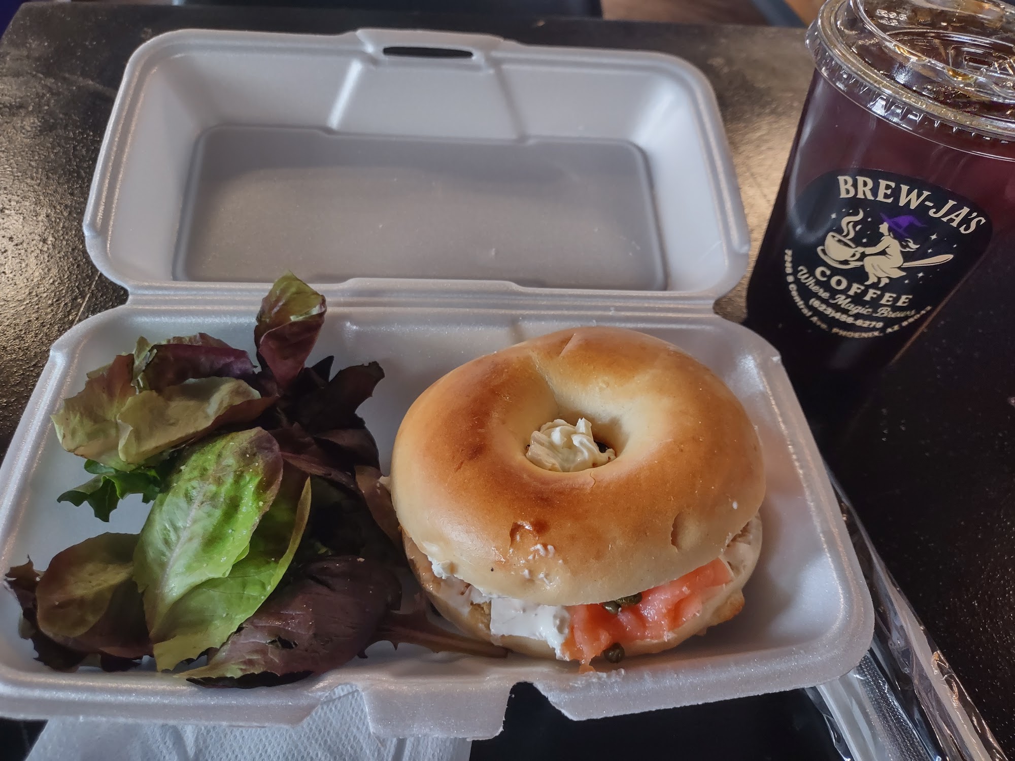 Lox Bagel in clamshell with mixed greens and branded dark iced coffee