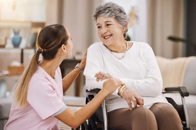 Une infirmière souriante s’agenouille à côté d’une femme âgée heureuse en fauteuil roulant, lui touchant doucement le bras et l’épaule.