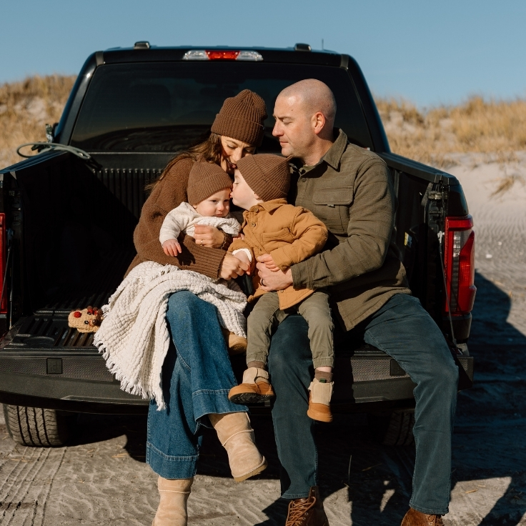 Intimate winter beach family moment with parents and their two young children cuddled together on a truck tailgate, wrapped in warm layers by the ocean.