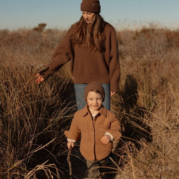 Mother following her young child as he walks through coastal dunes holding a stick, captured during a winter beach family session in warm neutral tones.