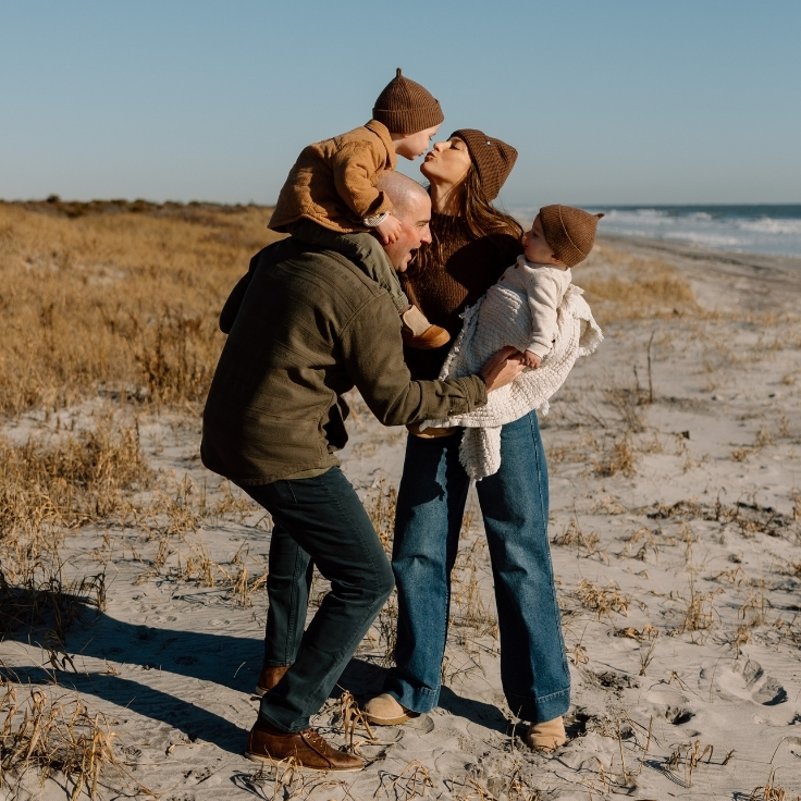Family of four bundled in neutral winter layers during a candid beach session, parents holding their young children with ocean and dunes in the background.