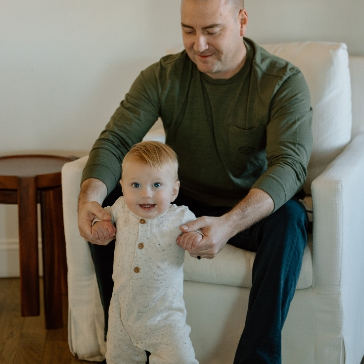 Father helping his baby stand in a cozy living room during an in-home family session, capturing early milestones and connection.