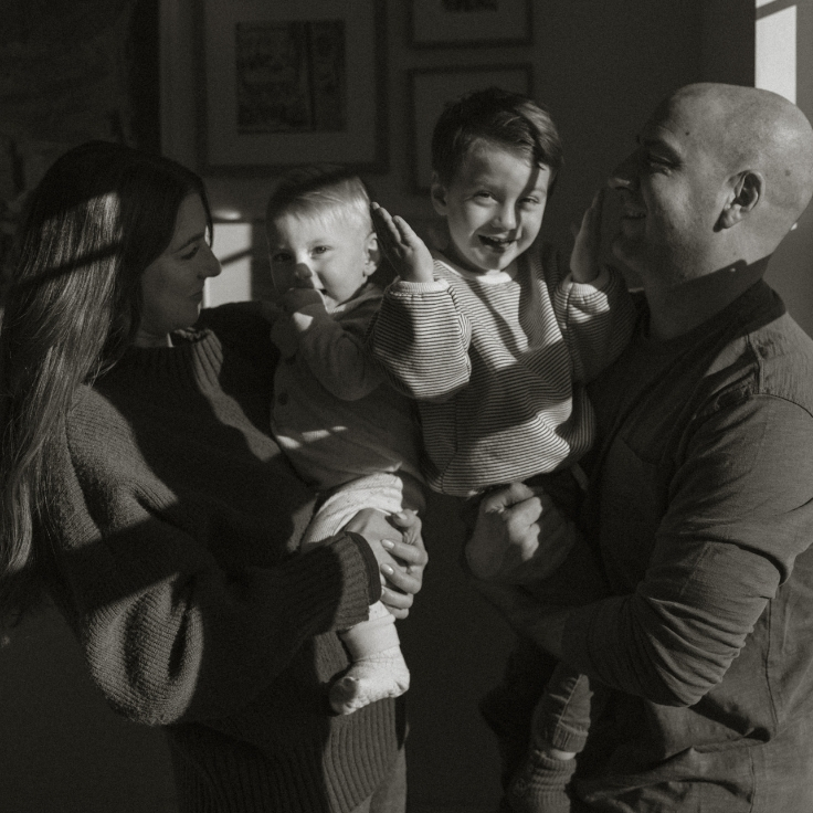 Black and white candid family moment at home, with parents holding their two young children in bright contrasted window light.
