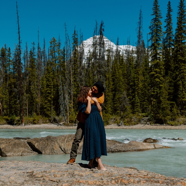 Couple embracing beside a glacial river surrounded by evergreen trees and mountain scenery during an adventurous engagement session in Yoho National Park, British Columbia.
