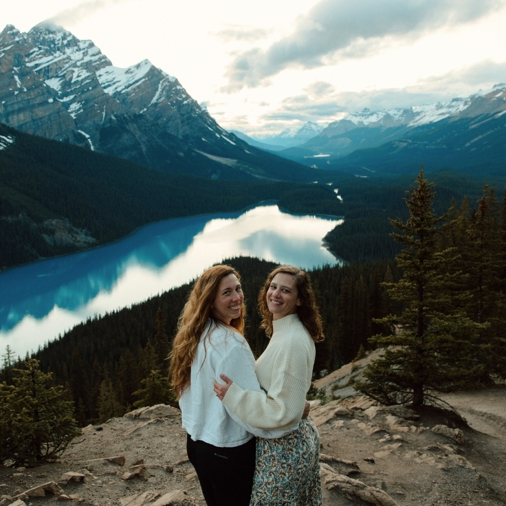 Photographer and bride smiling together at a scenic overlook above Peyto Lake with turquoise water, evergreen forest, and panoramic mountain views in Banff National Park.