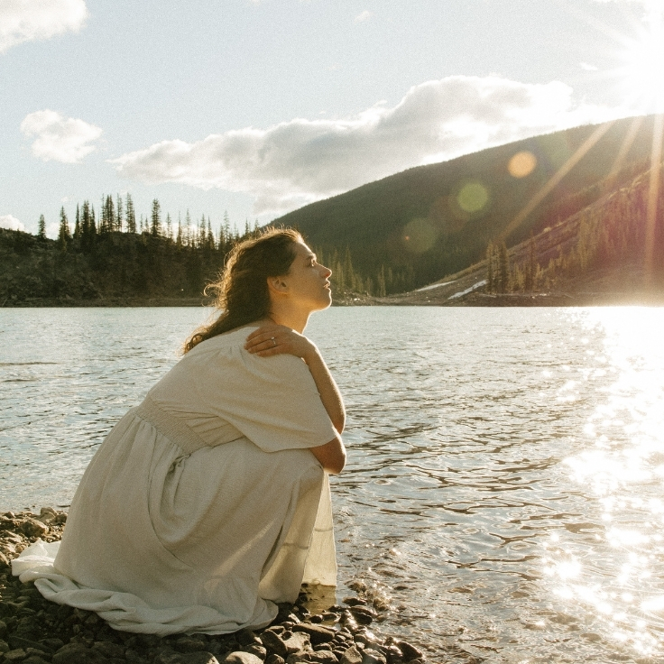 Bride-to-be sitting beside Moraine Lake during sunrise with glowing water reflections, mountain views, and cinematic golden light in Banff National Park.