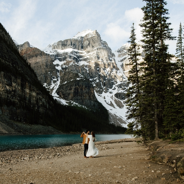 Couple standing beside Moraine Lake during a romantic Banff engagement session surrounded by turquoise water, towering mountains, and evergreen forest in the Canadian Rockies.
