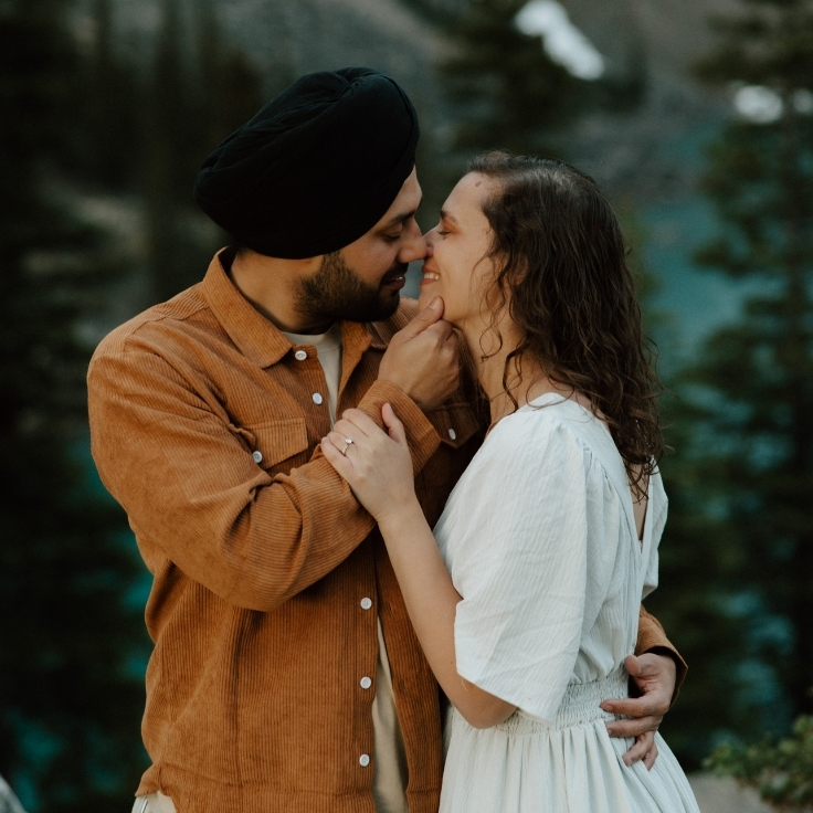 Engaged couple embracing and smiling beside a turquoise lake during a romantic Banff engagement session surrounded by evergreen trees and mountain scenery in the Canadian Rockies.