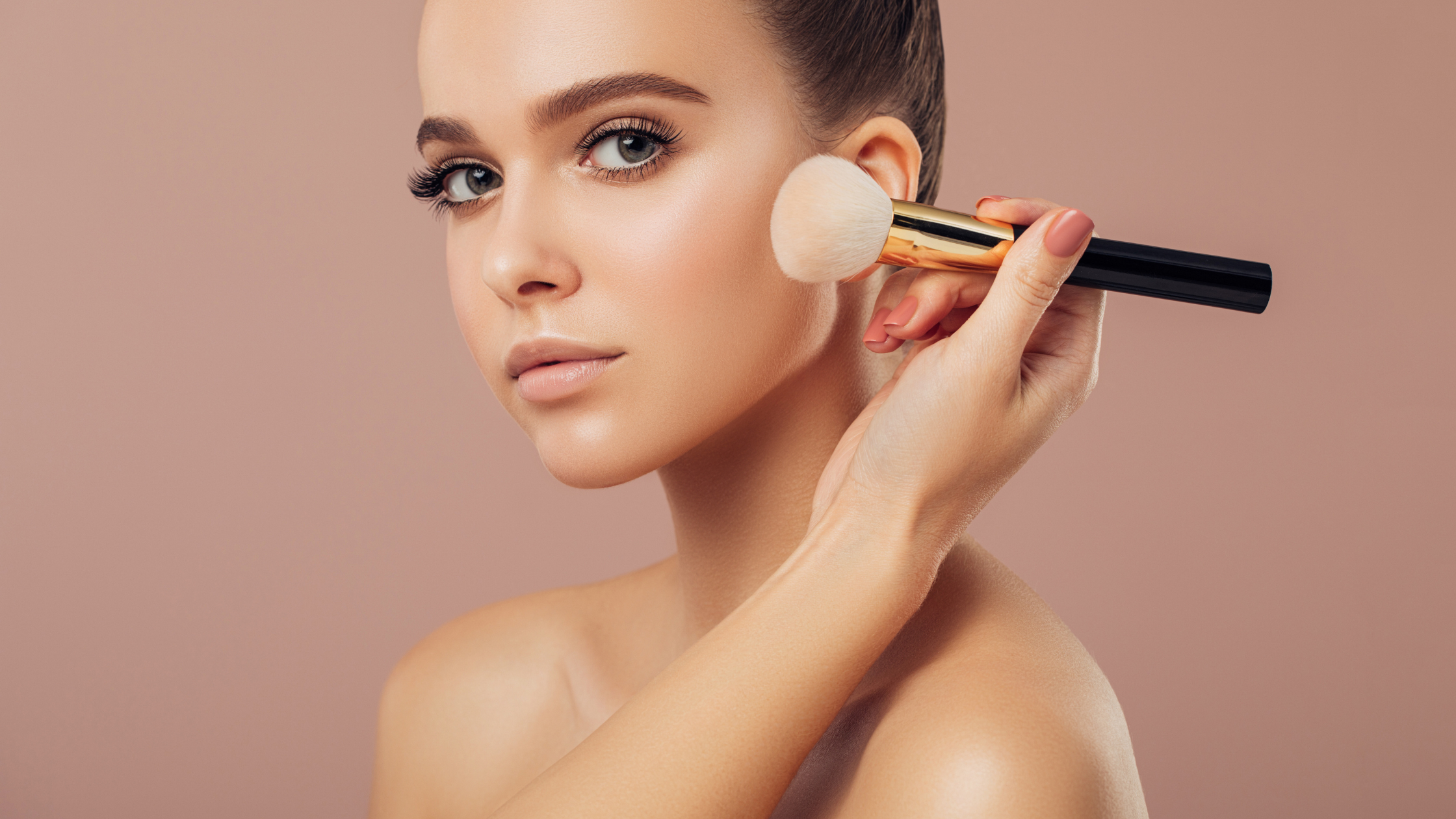 Close-up of a woman applying face powder with a makeup brush against a soft pink background. Close-up of a woman applying face powder with a makeup brush against a soft pink background.
