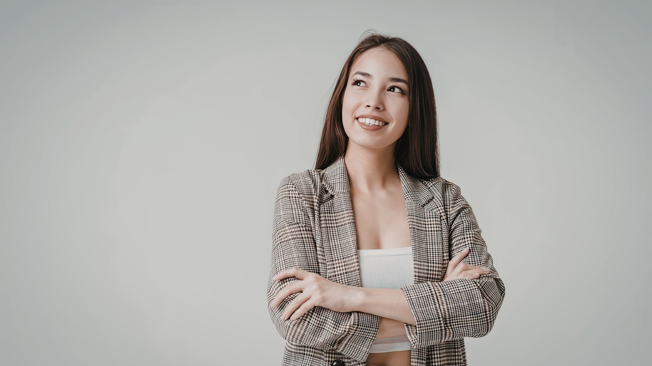 Young woman in a checked blazer standing with folded arms and smiling confidently against a plain light background. Young woman in a checked blazer standing with folded arms and smiling confidently against a plain light background.