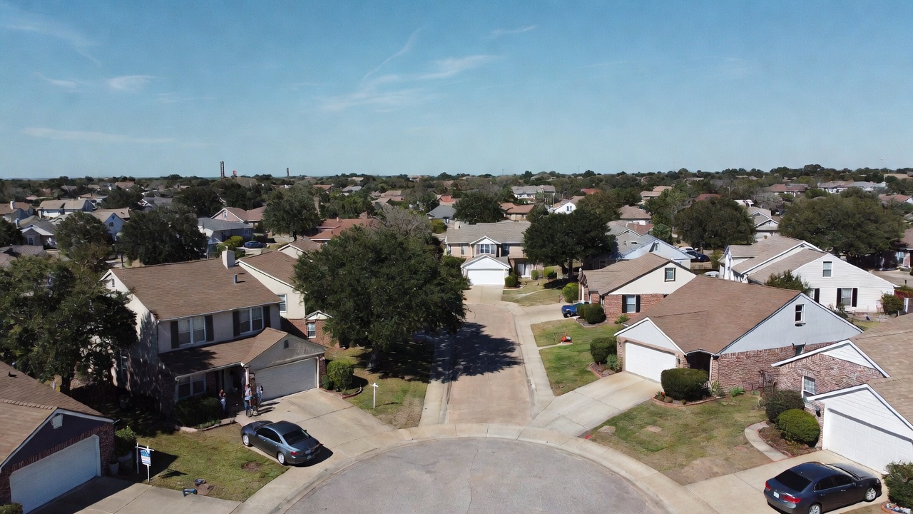 Drone aerial view of a suburban cul-de-sac with single-family homes, driveways, mature trees, and blue sky