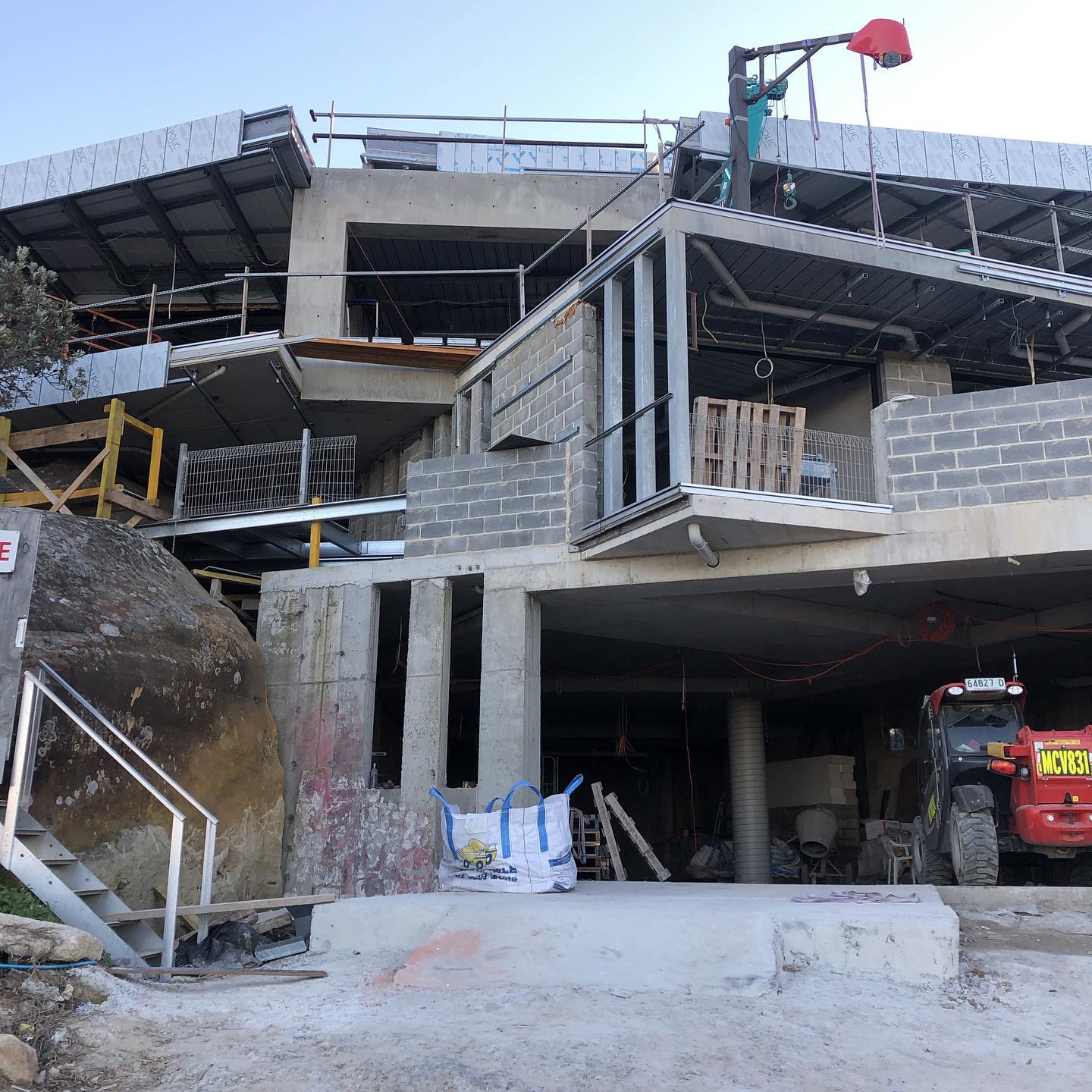 A construction site featuring a multi-level building under construction. The structure has exposed concrete, steel framing, and brickwork, with scaffolding and safety barriers in place. A large rock formation is visible on the left, and a construction vehicle is parked on the right near bags of building materials and tools.