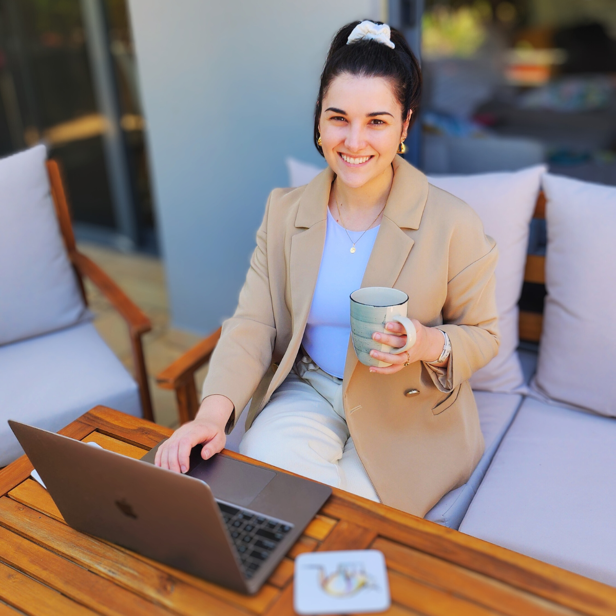 Aly Nicolaou sitting outdoors at a wooden table with a laptop, holding a coffee mug and smiling at the camera. She is dressed in a beige blazer and light-coloured outfit, appearing approachable and relaxed.